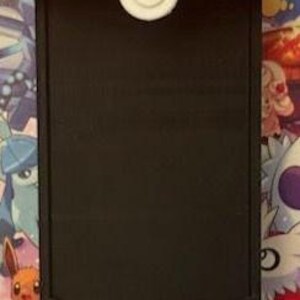 Pode incluir: Um suporte para telem&oacute;vel com o tema Pokeball, vermelho e branco. A parte superior &eacute; vermelha, o meio tem um espa&ccedil;o retangular preto e a parte inferior &eacute; branca. Um bot&atilde;o branco est&aacute; centrado na parte superior vermelha.