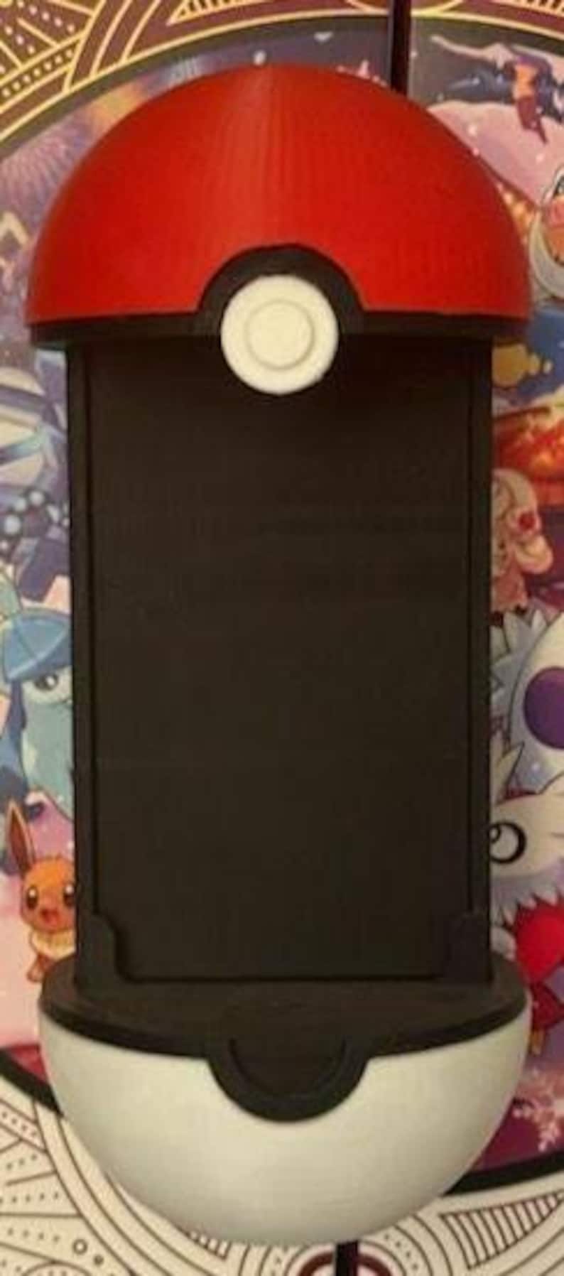 Pode incluir: Um suporte para telem&oacute;vel com o tema Pokeball, vermelho e branco. A parte superior &eacute; vermelha, o meio tem um espa&ccedil;o retangular preto e a parte inferior &eacute; branca. Um bot&atilde;o branco est&aacute; centrado na parte superior vermelha.