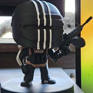 May include: A black figurine of a character wearing a helmet with three white stripes and holding a gun. The figurine has the word "KILLA" on the chest. The figure is on a black circular base.