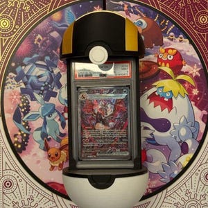 Op de afbeelding: Een zwart, wit en goudkleurig display in de vorm van een Pok&eacute; Ball, met een beoordeelde ruilkaart. De kaart bevat een kleurrijke illustratie van een personage. De achtergrond is een decoratief paneel met Pok&eacute;mon-personages.