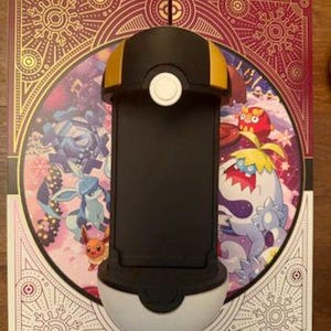 Op de afbeelding: Een zwart-witte telefoonhouder met gouden accenten, ontworpen om op een Pok&eacute; Ball te lijken. De houder staat op een decoratieve achtergrond met kleurrijke cartoonfiguren en sierlijke patronen. De telefoonhouder is ontworpen om een telefoon vast te houden.