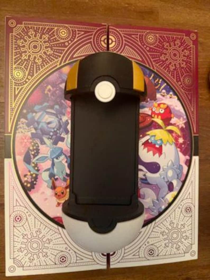 Op de afbeelding: Een zwart-witte telefoonhouder met gouden accenten, ontworpen om op een Pok&eacute; Ball te lijken. De houder staat op een decoratieve achtergrond met kleurrijke cartoonfiguren en sierlijke patronen. De telefoonhouder is ontworpen om een telefoon vast te houden.