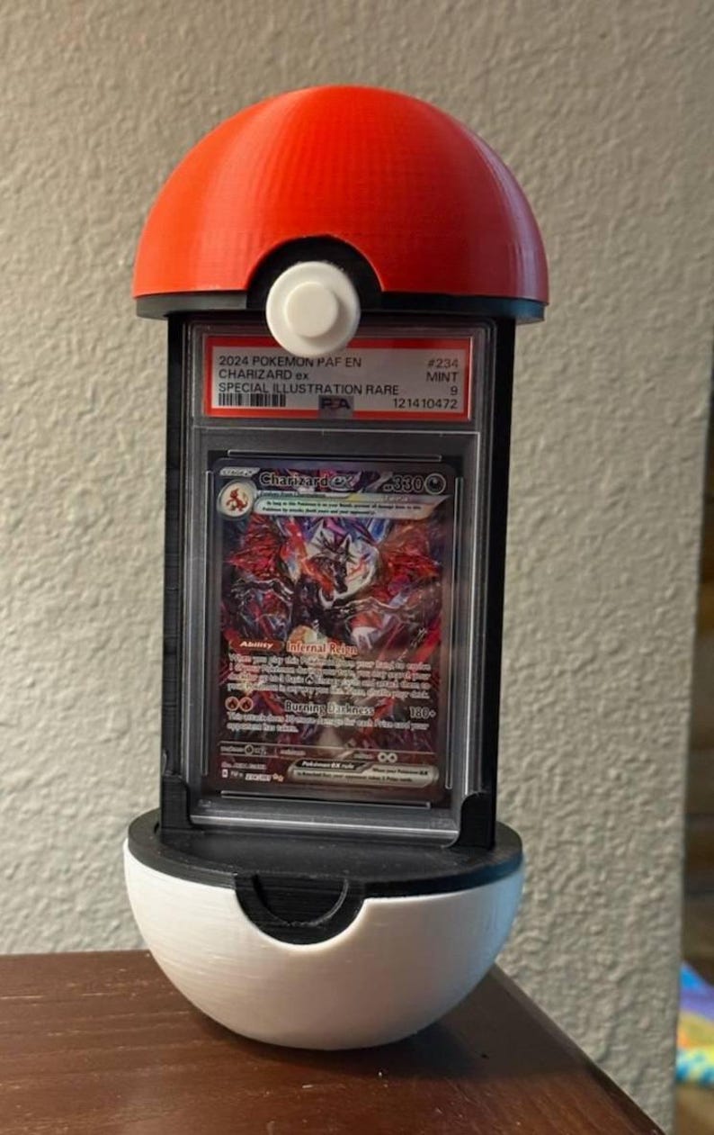 May include: A red and white 3D-printed Poké Ball display case holding a graded Pokémon card. The card features a Charizard ex illustration, with text indicating it's a Special Illustration Rare from 2024.