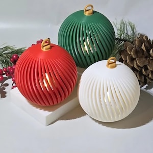 Könnte beinhalten: Drei dekorative, kugelförmige Ornamente in Rot, Grün und Weiß. Jedes Ornament hat ein Spiraldesign und eine goldfarbene Schlaufe oben. Die Ornamente sind von innen beleuchtet und erzeugen ein warmes Leuchten. Festliche Weihnachtsdekoration.