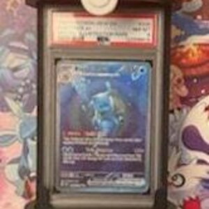 Pode incluir: Um suporte de exibi&ccedil;&atilde;o vermelho e branco em forma de Pok&eacute; Bola, segurando um cart&atilde;o colecion&aacute;vel classificado. O cart&atilde;o apresenta uma ilustra&ccedil;&atilde;o azul e branca de uma criatura. O suporte tem uma moldura preta e um detalhe de bot&atilde;o branco.