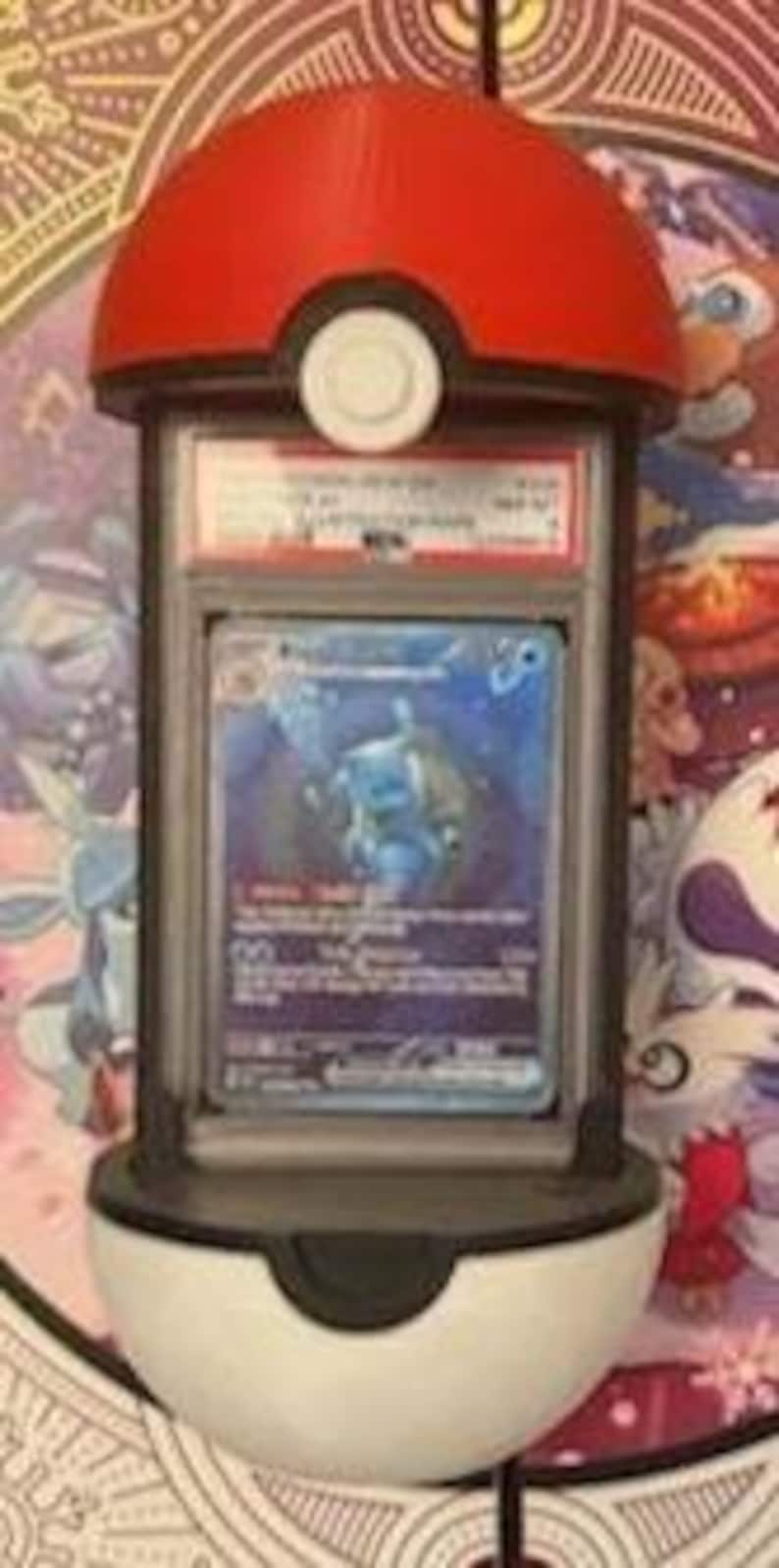 Pode incluir: Um suporte de exibi&ccedil;&atilde;o vermelho e branco em forma de Pok&eacute; Bola, segurando um cart&atilde;o colecion&aacute;vel classificado. O cart&atilde;o apresenta uma ilustra&ccedil;&atilde;o azul e branca de uma criatura. O suporte tem uma moldura preta e um detalhe de bot&atilde;o branco.