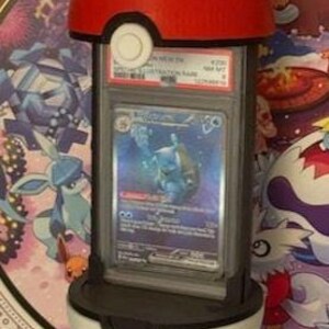 Pode incluir: Um suporte de exibi&ccedil;&atilde;o de Pok&eacute; Bola vermelho e branco segurando uma carta colecion&aacute;vel Pok&eacute;mon classificada. O suporte &eacute; projetado para se assemelhar a uma Pok&eacute; Bola, com um topo vermelho e uma parte inferior branca. O cart&atilde;o est&aacute; em um suporte de prote&ccedil;&atilde;o.