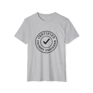 May include: A heather gray t-shirt featuring a circular graphic. The graphic reads "CERTIFIED PROMPT ENGINEER" around a checkmark symbol. The shirt has short sleeves and a crew neck.