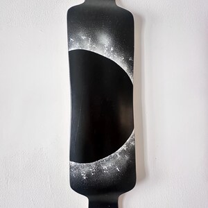 May include: Black longboard with a unique design. The board features a large black crescent shape with white speckled accents. The board is a solid black color with a rounded top and a curved bottom. The board is hanging on a white wall.
