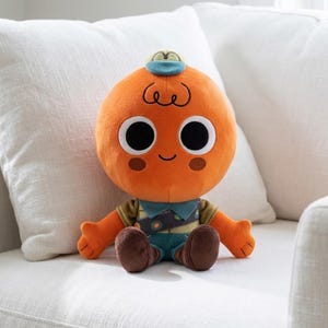 May include: A plush toy featuring a round orange head with large black eyes, a small smile, and brown circular cheeks. The character wears a small blue cap, a tan shirt with a blue collar, a sash with circular badges, and dark brown pants.