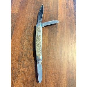 以下が含まれることがあります： 3枚の刃が付いたヴィンテージのポケットナイフ。そのうち2枚が開いています。ハンドルは、テクスチャード加工されたパターンとシルバーカラーの楕円形のアクセントが付いた、明るいベージュ色です。ブレードはシルバーで、ナイフは木製の表面にあります。