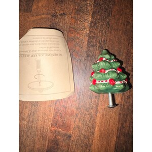 Puede incluir: Un pomo de cerámica verde con forma de árbol de Navidad con detalles de bayas rojas y un tornillo plateado. El pomo está diseñado para muebles o armarios. Un papel con instrucciones está al lado del pomo.