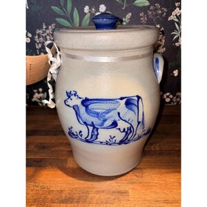 May include: A light gray stoneware crock with a blue painted cow illustration on the front. The cylindrical container features two side handles, a matching lid with a blue knob, and a tan paper tag attached to the side with a white ribbon.