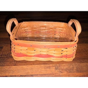 May include: A rectangular woven basket with a clear glass insert and two arched handles. The basket features alternating bands of orange and red, with a decorative red band around the top. The handles are tan.