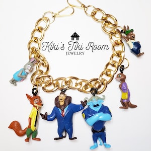 Puede incluir: Una pulsera de cadena dorada con dijes de personajes de dibujos animados. La pulsera presenta una cadena de eslabones grandes y un cierre de mosquet&oacute;n. Los dijes incluyen un zorro, un le&oacute;n, un conejo, una nutria y un b&uacute;falo azul. Tambi&eacute;n se ve el texto "Kiki's Tiki Room Jewelry".