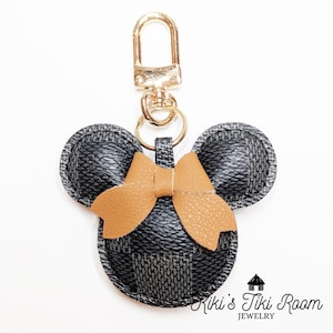 Könnte beinhalten: Ein schwarz-grauer, karierter Leder-Schlüsselanhänger in Form eines Mäusekopfes mit einer beigefarbenen Schleife. Der Schlüsselanhänger hat einen goldfarbenen Verschluss und Ring. Das Bild enthält den Text "Kiki's Tiki Room JEWELRY."