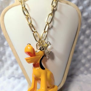 Puede incluir: Un collar de cadena dorada con un colgante de perro amarillo que tiene un collar rojo y la boca abierta. El colgante está decorado con un pequeño dije de perla blanca y cristales transparentes, suspendido de una cadena de eslabones ovalados.