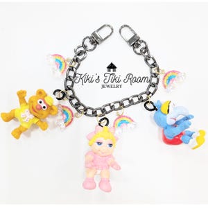 Peut inclure: Un bracelet à chaîne en métal foncé avec trois breloques représentant des personnages Muppet : un ours jaune, un cochon rose et une créature bleue. Le bracelet est orné de petites breloques arc-en-ciel scintillantes et porte le texte Kiki's Tiki Room JEWELRY.
