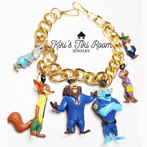 Puede incluir: Pulsera de cadena dorada con dijes de personajes de dibujos animados. Los dijes incluyen un zorro, un le&oacute;n y otras figuras de animales. La pulsera tiene un cierre de mosquet&oacute;n. El texto "Kiki's Tiki Room Jewelry" es visible.