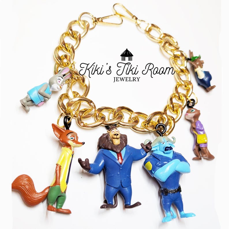 Puede incluir: Pulsera de cadena dorada con dijes de personajes de dibujos animados. Los dijes incluyen un zorro, un le&oacute;n y otras figuras de animales. La pulsera tiene un cierre de mosquet&oacute;n. El texto "Kiki's Tiki Room Jewelry" es visible.