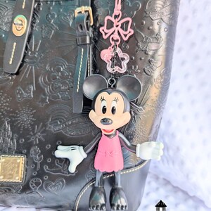 May include: A Minnie Mouse keyring with a pink dress and bow charm hangs from a black handbag. The handbag has embossed designs and gold hardware. A pink bow and star charm dangle above Minnie.