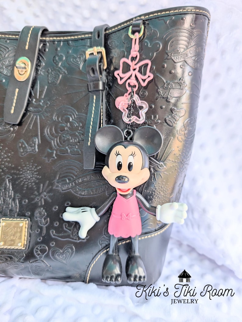 May include: A Minnie Mouse keyring with a pink dress and bow charm hangs from a black handbag. The handbag has embossed designs and gold hardware. A pink bow and star charm dangle above Minnie.