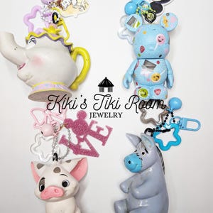 May include: Collection of character keychains, including a white teapot with yellow accents, a blue Mickey Mouse-inspired figure, a pink pig, and a gray donkey. Each keychain has a unique design and color scheme, perfect for Disney fans.
