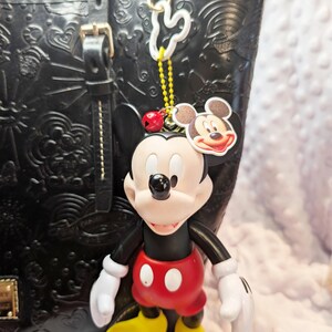 May include: A Mickey Mouse keyring in red, yellow, black, and white hangs from a black leather tote bag. The keyring features a Mickey Mouse figure and a Mickey Mouse head tag. The bag has embossed designs.