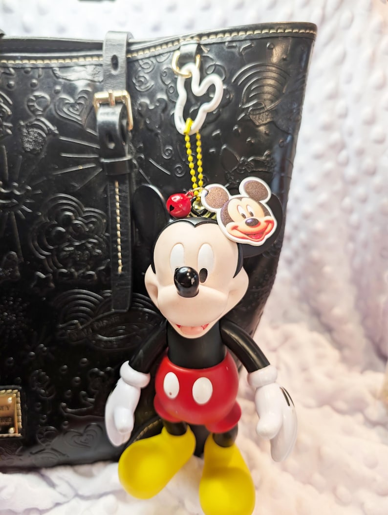 May include: A Mickey Mouse keyring in red, yellow, black, and white hangs from a black leather tote bag. The keyring features a Mickey Mouse figure and a Mickey Mouse head tag. The bag has embossed designs.