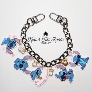 May include: A silver-toned chain bracelet with lobster clasp closures, adorned with cartoon Stitch charms in various poses. The charms are blue and white, with a heart-shaped charm and the text "Kiki's Tiki Room JEWELRY".