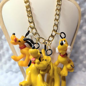 May include: A gold-tone metal chain necklace featuring three yellow dog character charms. Each charm depicts a cartoon dog with black ears, a red collar, and distinct poses. The charms are attached to the chain with black metal jump rings.