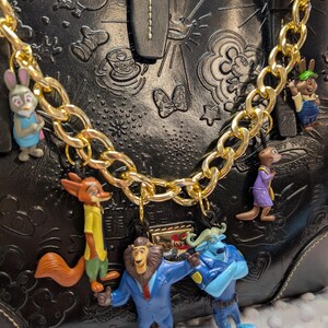 Puede incluir: Bolso de mano de cuero negro con dise&ntilde;os Disney en relieve, adornado con una cadena dorada y colgantes de personajes de la pel&iacute;cula Zootr&oacute;polis. Los colgantes incluyen a Judy Hopps, Nick Wilde y otros personajes. El bolso tiene un cierre dorado.