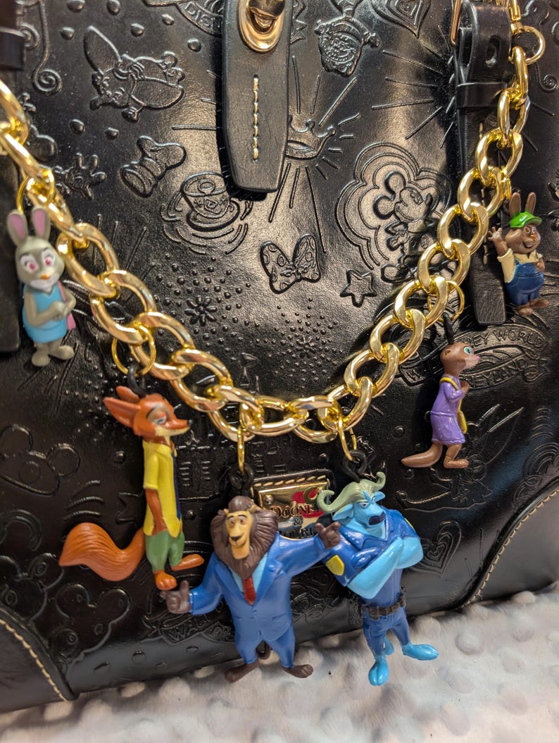 Puede incluir: Bolso de mano de cuero negro con dise&ntilde;os Disney en relieve, adornado con una cadena dorada y colgantes de personajes de la pel&iacute;cula Zootr&oacute;polis. Los colgantes incluyen a Judy Hopps, Nick Wilde y otros personajes. El bolso tiene un cierre dorado.