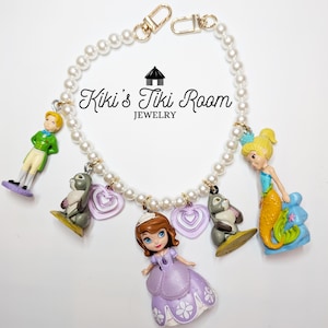 Peut inclure: Un bracelet de perles avec des fermoirs et des breloques dorés. Les breloques comprennent un prince, une sirène, une princesse, deux lapins et deux breloques en forme de cœur violet. Le texte "Kiki's Tiki Room Jewelry" est au centre.