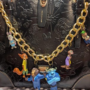 Puede incluir: Bolso de mano de cuero negro con cadena dorada y abalorios de personajes de Disney. El bolso presenta dise&ntilde;os en relieve de Disney y un cierre dorado. Los abalorios incluyen personajes de Zootopia y otras pel&iacute;culas de Disney.
