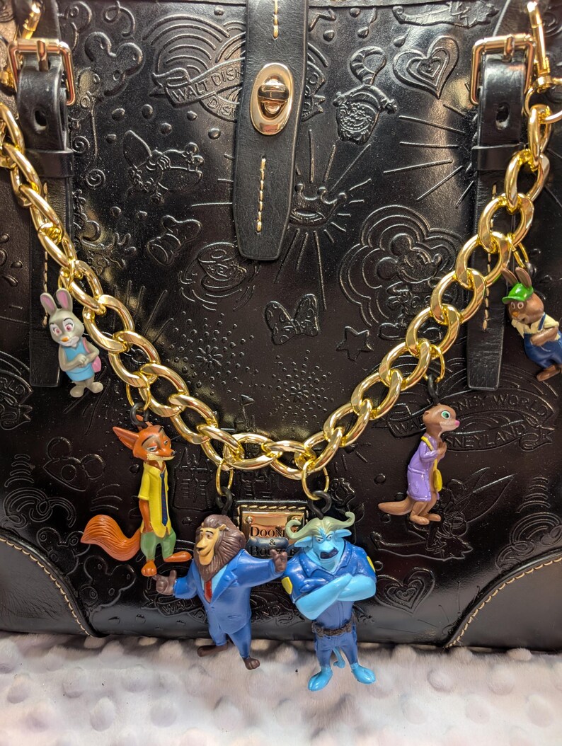 Puede incluir: Bolso de mano de cuero negro con cadena dorada y abalorios de personajes de Disney. El bolso presenta dise&ntilde;os en relieve de Disney y un cierre dorado. Los abalorios incluyen personajes de Zootopia y otras pel&iacute;culas de Disney.