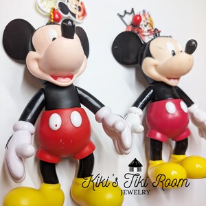May include: Two Mickey Mouse figurines. Each figure has a black torso and arms, red shorts, white gloves, and yellow shoes. The figures are holding hands. The background is white. The text "Kiki's Tiki Room Jewelry" is at the bottom.