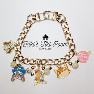 Puede incluir: Pulsera de dijes dorada con dijes de esmalte coloridos. Los dijes incluyen una placa de policía, un conejito, un zorro, una flor y un par de personajes. La pulsera tiene un diseño de cadena gruesa y un cierre de broche. También se ve el texto "Kiki's Tiki Room JEWELRY".