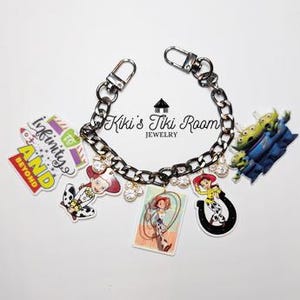 May include: A silver-toned chain bracelet with various Toy Story-themed charms. Charms include "Infinity and Beyond" text, a cowgirl, a horseshoe, and aliens. The bracelet also features clear, sparkling accents. The text "Kiki's Tiki Room Jewelry" is visible.