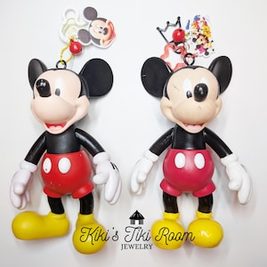 May include: Two Mickey Mouse figurines. Each figure has a black body, red shorts, white gloves, and yellow shoes. One has a Mickey Mouse head charm, and the other has a charm with multiple Disney characters. The text "Kiki's Tiki Room Jewelry" is at the bottom.