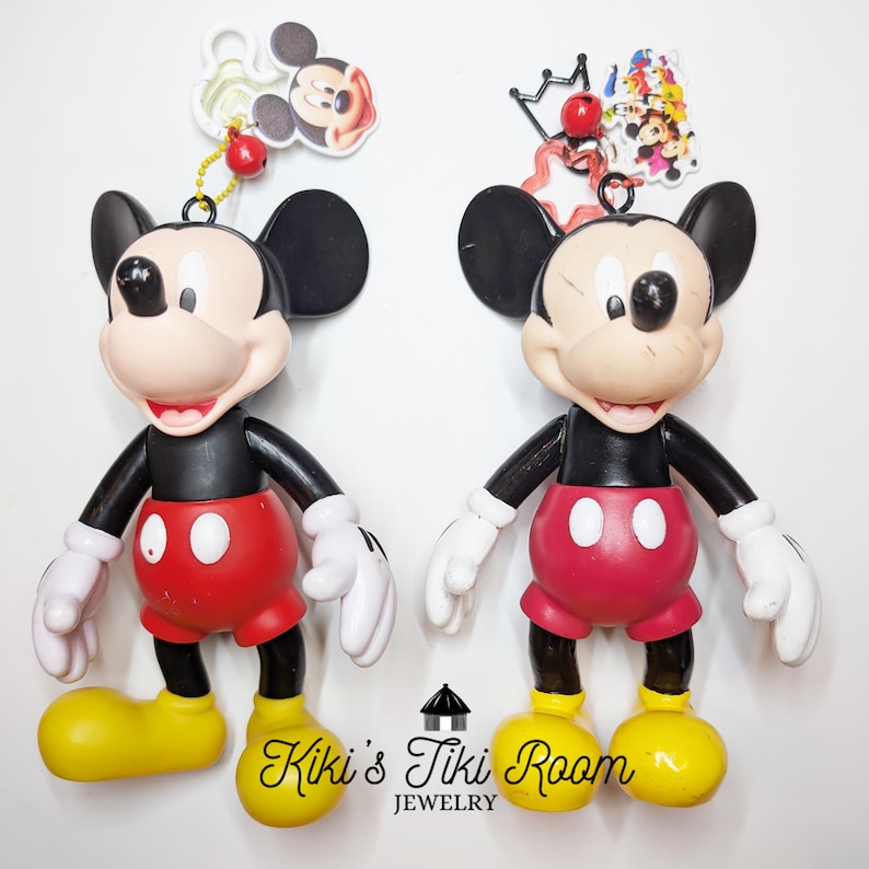 May include: Two Mickey Mouse figurines. Each figure has a black body, red shorts, white gloves, and yellow shoes. One has a Mickey Mouse head charm, and the other has a charm with multiple Disney characters. The text "Kiki's Tiki Room Jewelry" is at the bottom.