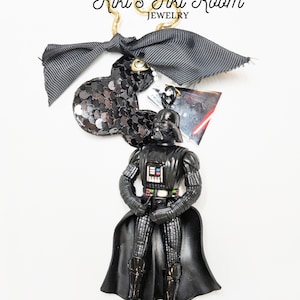 Puede incluir: Un llavero con una figura negra de Darth Vader con una cabeza de Mickey Mouse con lentejuelas negras y un lazo a rayas grises. El llavero está sujeto a un anillo de metal dorado. El texto "Kiki's Tiki Room JEWELRY" está en la parte superior.