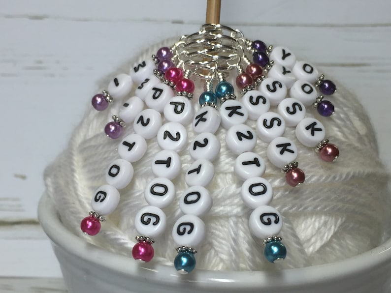 May include: White plastic stitch markers with black numbers and letters, including "K2tog", "SSK", "YO", "K", "P2tog", "1tog", "2tog", "G", "O", "1", "2", "3", and "4". The markers are attached to a silver ring and have colorful beads.