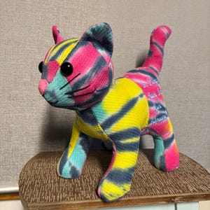 May include: A colorful tie-dye cat plush toy with black button eyes, a pink nose, and a pink and blue striped tail. The cat is standing on a small wooden table. The fabric has a mix of pink, yellow, blue, and black colors.