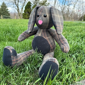 May include: A handmade plush bunny toy with a plaid fabric pattern in shades of brown and black. The bunny has a black nose and pink felt nose. The bunny is sitting on a green grassy surface.