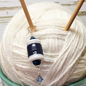 May include: A white yarn ball with two wooden knitting needles sticking out of it. A blue and white yarn counter with a crystal bead hangs from the needles.