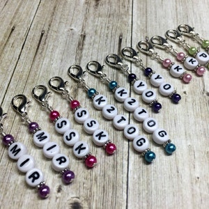May include: Ten colourful stitch markers with white beads and silver clips. Each marker has a different letter or number on it, including "MIR", "SSK", "K2TOG", "YO", "K", and "P".