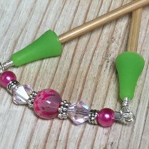 May include: A pair of wooden knitting needles with green silicone tips and a pink and silver beaded charm attached to the needles.