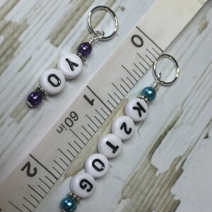 May include: Two white plastic stitch markers with silver rings and beads. The first marker has purple beads and the letters "Y" and "O". The second marker has blue beads and the numbers "6", "0", "1", and "2".