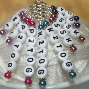 May include: White plastic stitch markers with black lettering, including numbers and letters, with colorful beads attached to each marker. The markers are arranged in a circle on a white yarn ball.